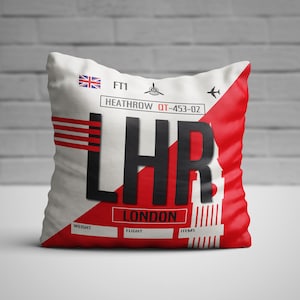 Puede incluir: Almohada blanca y roja con un diseño de etiqueta de equipaje de aeropuerto en blanco y negro. La etiqueta dice "Heathrow" y "LHR London" con una bandera británica roja, blanca y azul.