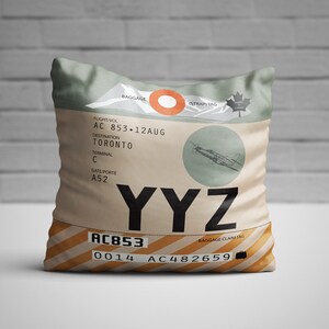 May include: A decorative pillow with a vintage luggage tag design. The pillow features the destination "TORONTO" and the airport code "YYZ". The design includes a stylized airplane illustration and the text "AC 853-12AUG". The pillow has a beige and green color scheme.