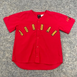 May include: A red button-up baseball jersey with the word "JORDAN" arched across the front in yellow lettering with black outlines. The sleeves have the word "JORDAN" printed on them. A Nike logo is visible on the tag.