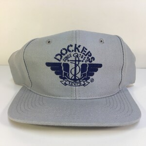 Peut inclure: Casquette de baseball grise avec un logo brodé bleu qui dit "Dockers Since 1850" avec une ancre et des ailes et le texte "Levi's" en dessous.