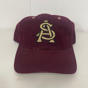 Op de afbeelding: Bordeauxrode baseballpet met een goudkleurig geborduurd "AS"-logo op de voorkant. De pet heeft een gebogen klep en een gestructureerde kroon. Twee kleine beige knopen bevinden zich bovenop de pet.