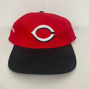 May include: A red baseball cap with a black brim. The cap features a white and red logo on the front. The word "HER" is embroidered on the side of the cap.