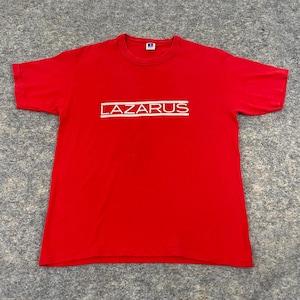 Könnte beinhalten: Ein rotes T-Shirt mit dem Wort "LAZARUS" in Weiß, horizontal zentriert gedruckt. Der Text ist in einer serifenlosen Schriftart und befindet sich innerhalb eines weißen rechteckigen Rahmens. Das T-Shirt ist aus weichem Material.