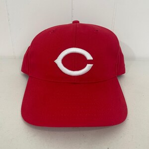 Pode incluir: Boné de beisebol vermelho com o logotipo "C" branco na frente. O boné tem uma aba curva e uma coroa arredondada. O chapéu é feito de um tecido liso, um acessório de equipe esportiva.