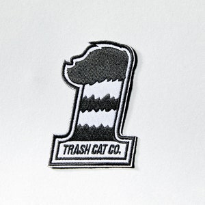 May include: Black and white embroidered patch featuring a cartoon raccoon wearing a striped hat with the text "TRASH CAT CO."
