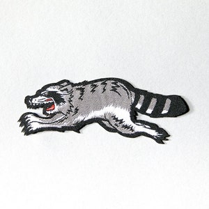May include: A black and white embroidered patch of a raccoon running with a striped tail.