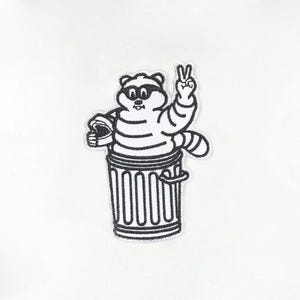 Op de afbeelding: Zwart-wit geborduurde patch met een cartoon wasbeer met een zonnebril en een gestreept shirt, die vanuit een vuilnisbak een vredesteken geeft. De patch heeft een witte achtergrond.