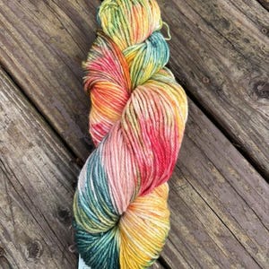 May include: A skein of colorful yarn in shades of yellow, orange, pink, green, and blue. The yarn is labeled with the yardage and the word "worsted". The yarn is displayed on a wooden surface.