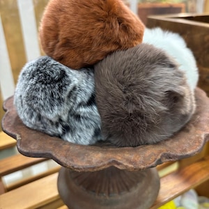 May include: A collection of fluffy, faux fur pom-poms in various colors, including brown, gray, and white, are stacked on a decorative, antique-style pedestal. The pom-poms have a soft, textured appearance, suggesting they are made from high-quality materials. The arrangement creates a visually appealing display.