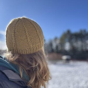 May include: A knitted, olive green beanie hat with vertical ribbing is worn by a person with long, wavy blonde hair. The person is facing away from the camera, with a bright blue sky and snowy background.