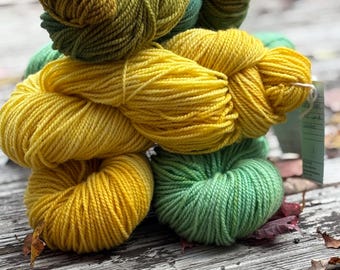 May include: A stack of multiple skeins of yarn in shades of bright yellow, golden yellow, and various tones of green. The yarn features a twisted, multi-ply texture and is bundled into rounded, soft shapes. A small rectangular tag is attached.