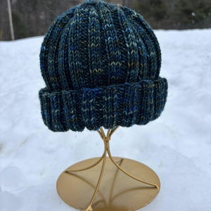 May include: A hand-knitted beanie hat in shades of blue and green, displayed on a gold-colored stand. The hat features a ribbed design and a folded brim. The hat is made of a soft, textured yarn, perfect for cold weather.
