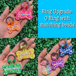 May include: A collection of colorful, bone-shaped pet tags with names spelled out in letter beads. Each tag has a silver ring and matching beads. The text "Ring Upgrade: O-Ring with matching beads" is displayed.