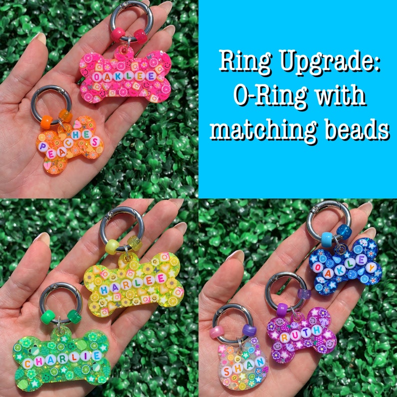 May include: A collection of colorful, bone-shaped pet tags with names spelled out in letter beads. Each tag has a silver ring and matching beads. The text "Ring Upgrade: O-Ring with matching beads" is displayed.