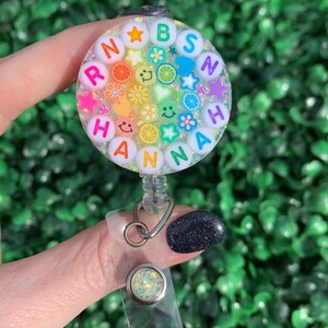 May include: A clear round badge holder with a rainbow of colorful letters spelling "RAINBOW" and "HANNAH" surrounded by glitter, stars, flowers, and smiley faces. The badge holder is attached to a clear plastic retractable badge reel.