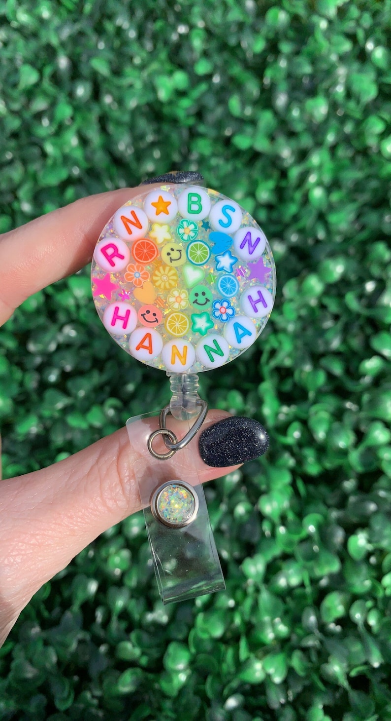 May include: A clear round badge holder with a rainbow of colorful letters spelling "RAINBOW" and "HANNAH" surrounded by glitter, stars, flowers, and smiley faces. The badge holder is attached to a clear plastic retractable badge reel.