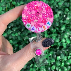 May include: Pink and white badge holder with the name "HALEY" spelled out in colorful beads. The badge holder has a pink glitter design with hearts, flowers, and smiley faces.
