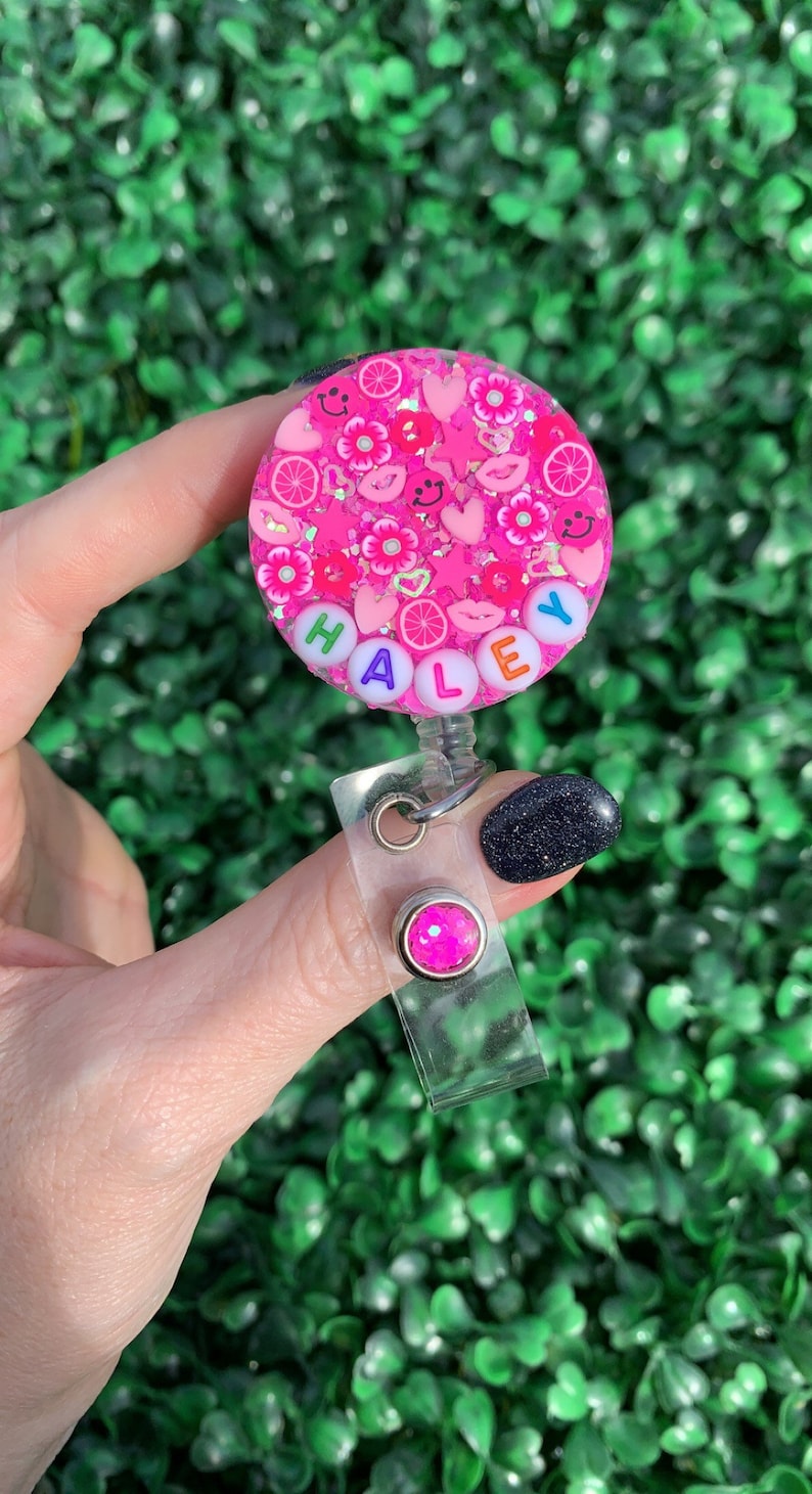 May include: Pink and white badge holder with the name "HALEY" spelled out in colorful beads. The badge holder has a pink glitter design with hearts, flowers, and smiley faces.