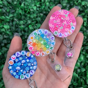 May include: Three colorful badge reels with clear plastic lanyards. The badge reels are decorated with glitter, small beads, and letters spelling out the names "Blake RVT", "Hannah BSN", and "Haley".