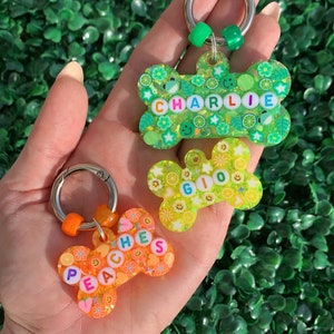 May include: Three colorful, decorative pet tags in the shape of bones and a flower. The tags are green, yellow, and orange, with the names "Charlie", "Gio", and "Peaches" spelled out in colorful beads. Each tag has a silver keyring.