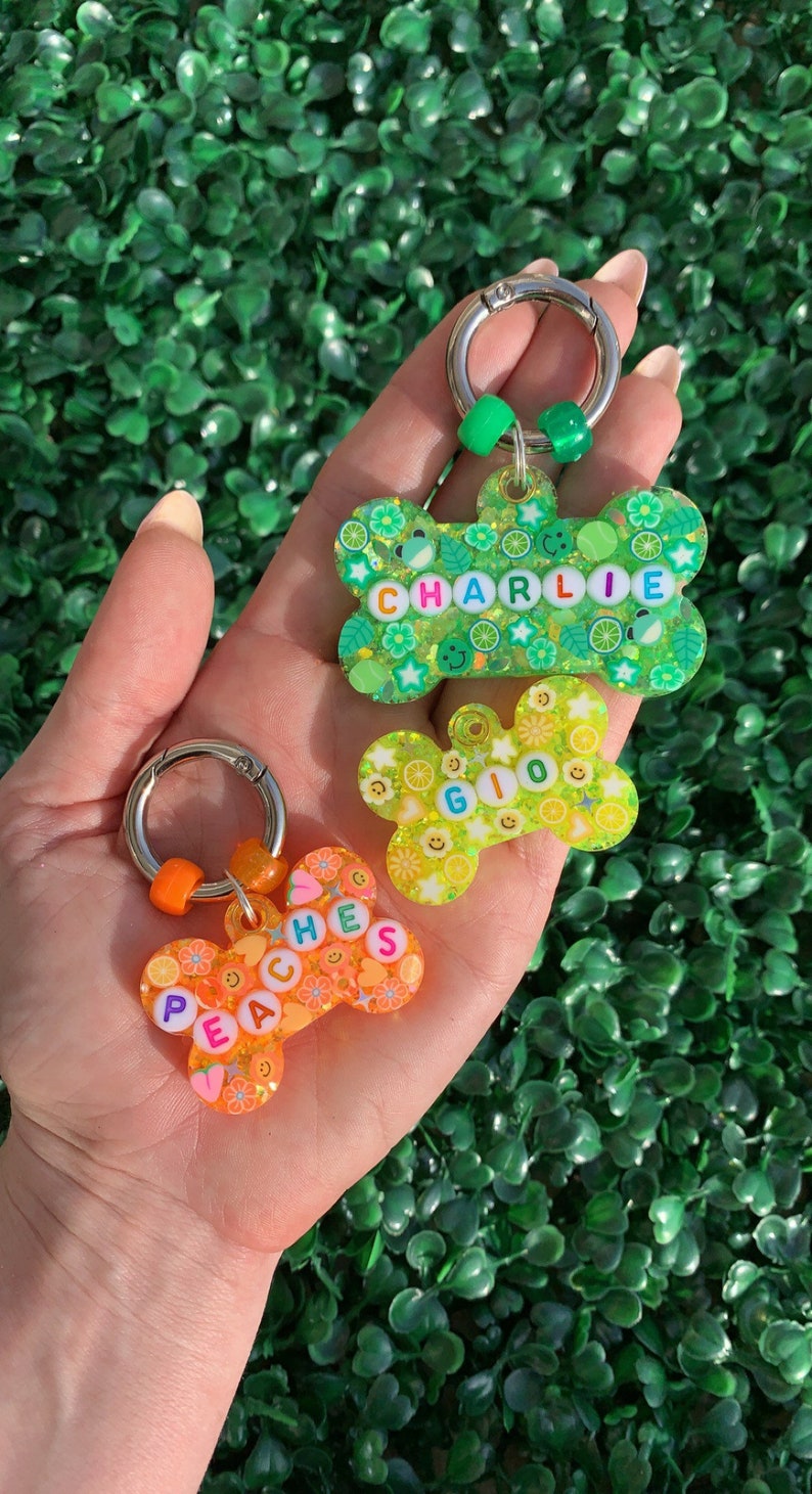 May include: Three colorful, decorative pet tags in the shape of bones and a flower. The tags are green, yellow, and orange, with the names "Charlie", "Gio", and "Peaches" spelled out in colorful beads. Each tag has a silver keyring.