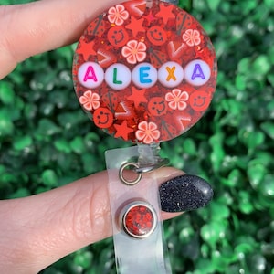May include: Red glitter badge holder with a clear plastic strap and a red glitter charm. The badge holder has the name "ALEXA" spelled out in white letters with colorful accents.