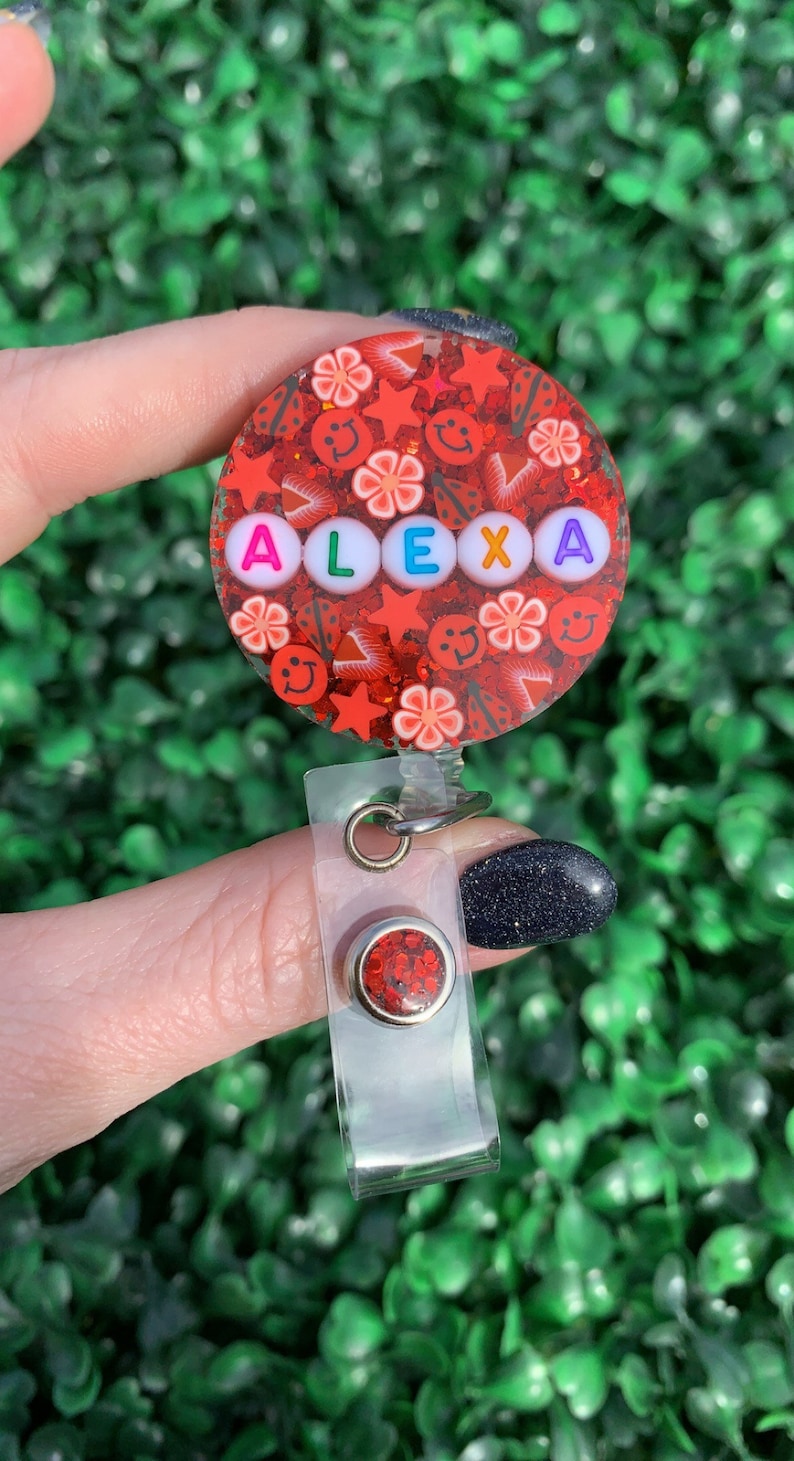 May include: Red glitter badge holder with a clear plastic strap and a red glitter charm. The badge holder has the name "ALEXA" spelled out in white letters with colorful accents.