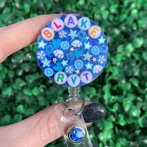 May include: A blue and white round badge holder with the name "BLAKE" spelled out in colorful letters. The badge holder is decorated with glitter, stars, hearts, clouds, and flowers. It is attached to a clear plastic retractable badge reel with a blue rhinestone.