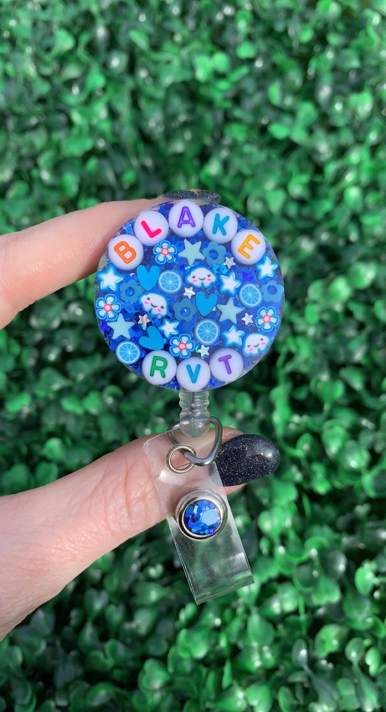 May include: A blue and white round badge holder with the name "BLAKE" spelled out in colorful letters. The badge holder is decorated with glitter, stars, hearts, clouds, and flowers. It is attached to a clear plastic retractable badge reel with a blue rhinestone.