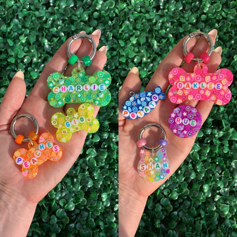 May include: A collection of colorful pet ID tags in various shapes, including bone and fish designs. The tags feature names like "CHARLIE," "GIO," "PEACHES," "OAKLEE," "RUE," and "SHAN" spelled out with letter beads, and are adorned with fruit and flower patterns.