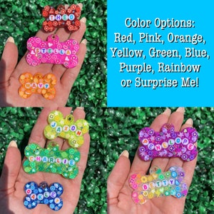 May include: A collection of colorful, bone-shaped pet tags with names spelled out in letter beads. The tags feature various designs and colors, including red, pink, orange, yellow, green, blue, purple, and rainbow. Text on the image lists color options.