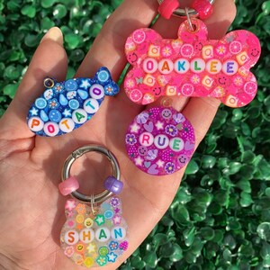 May include: A collection of colorful, handmade keychains in various shapes, including a bone, a fish, a circle, and a cat. Each keychain features a name spelled out with colorful letter beads and decorative elements like flowers and fruit slices. The keychains are held in a hand.