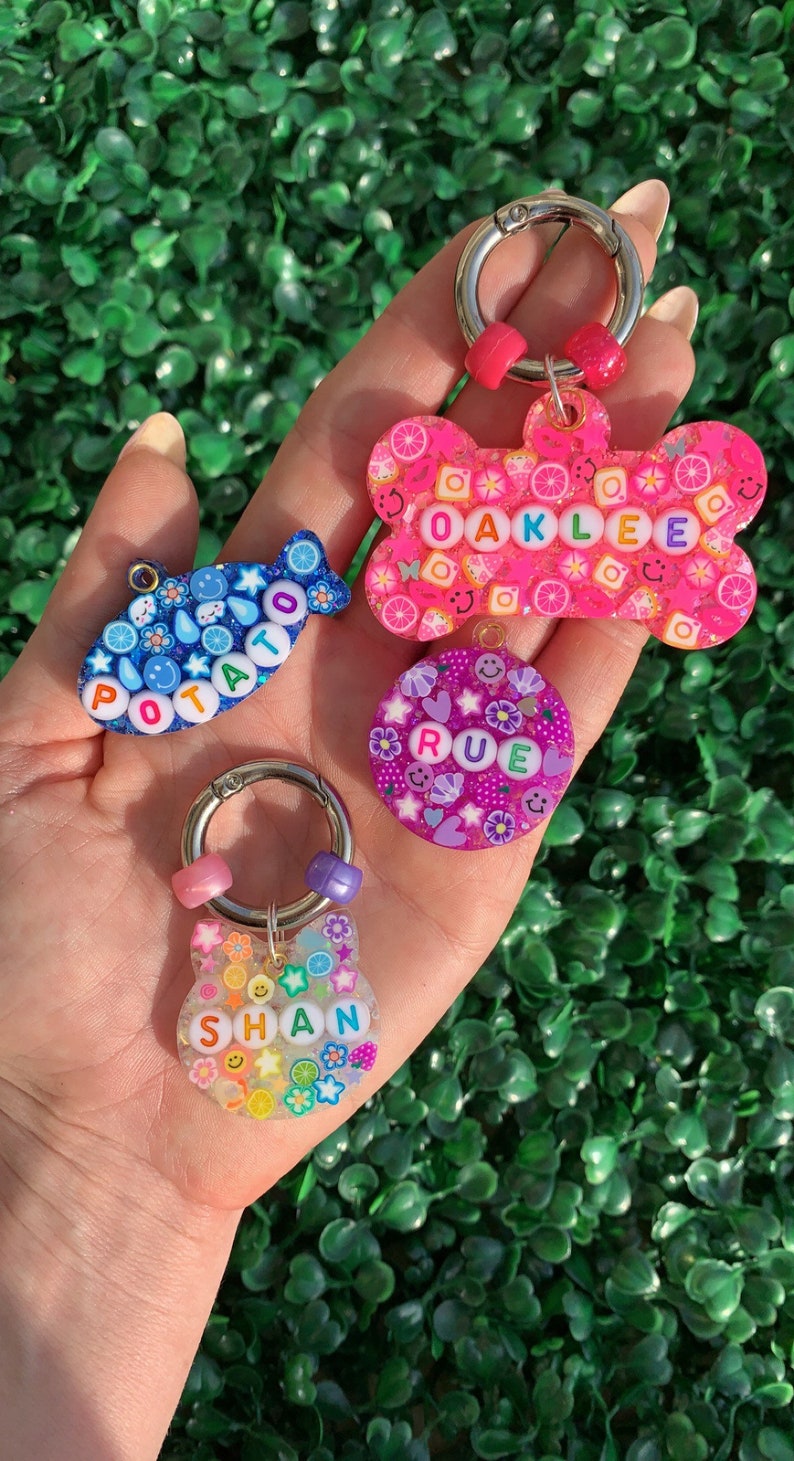 May include: A collection of colorful, handmade keychains in various shapes, including a bone, a fish, a circle, and a cat. Each keychain features a name spelled out with colorful letter beads and decorative elements like flowers and fruit slices. The keychains are held in a hand.