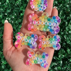 May include: A collection of colorful, personalized pet tags in various shapes. Each tag features a name spelled out with letter beads and is decorated with small, cheerful embellishments like smiley faces and flowers. The tags are held against a backdrop of green foliage.