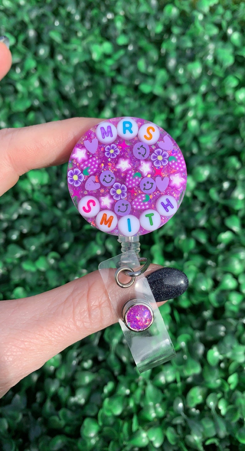 May include: Purple glitter badge holder with the name "MRS SMITH" spelled out in colorful letters. The badge holder has a clear plastic strap and a small purple glitter charm.