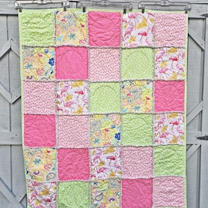 May include: A patchwork quilt with a variety of fabrics in pink, green, and yellow. The quilt features squares with flamingo patterns, polka dots, and floral designs. The quilt is hanging on a clothesline.