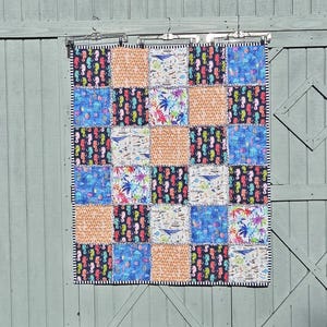 May include: A patchwork quilt with a variety of patterned squares, including seahorses, palm trees, and nautical themes. The quilt features a black and white striped border and is displayed on a weathered wooden door.