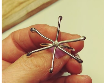 May include: A silver-colored ring with a starburst design. The ring features six prongs extending outwards from a central point, each ending in a rounded tip. The ring is held up by a person's hand.
