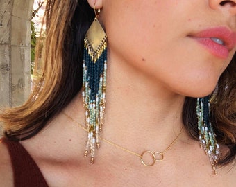 May include: A pair of long, beaded earrings with a gold geometric shape at the top. The earrings are made with a gradient of green, blue, and white beads. The earrings are worn by a person with long brown hair.
