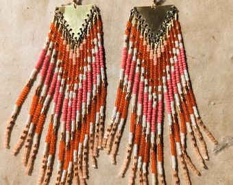 May include: A pair of long, beaded earrings with a geometric design. The earrings are made with orange, pink, and white beads and have a gold-toned metal top.