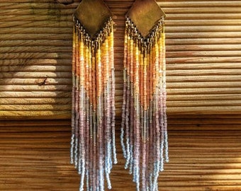 May include: Long, beaded earrings with a geometric top and a cascade of colourful beads in shades of yellow, peach, pink, and blue. The earrings are displayed against a wooden background, with a pink and purple flower at the top.