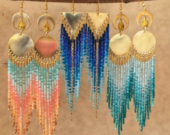 May include: Three pairs of gold tone dangle earrings with beaded fringe. The earrings feature a crescent moon and a circle with a sunburst design. The fringe is ombre, with shades of pink, blue, and turquoise.
