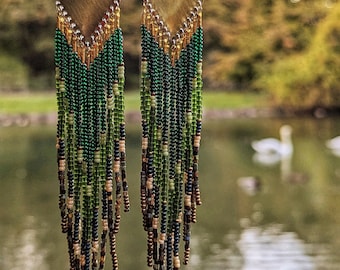 May include: A pair of long, green and gold beaded earrings with a brass triangle pendant. The earrings are hanging from a branch over a pond.