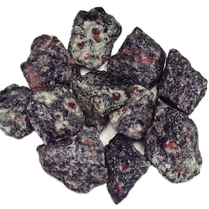 May include: A group of small, rough, dark gray stones with red ruby inclusions. The stones are clustered together on a white background.
