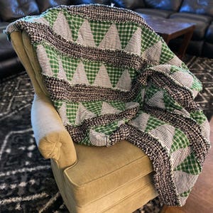 May include: A green and black patchwork quilt with a triangle pattern. The quilt is made of various fabrics, including plaid and striped fabrics. The quilt is draped over a chair.
