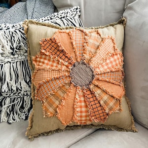 Może przedstawiać: Dekoracyjna poduszka z dużym wzorem pomarańczowego i brązowego kwiatu. Poduszka jest wykonana z patchworku tkanin i ma strzępiony brzeg.