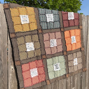 Op de afbeelding: Een patchworkquilt met negen vierkanten van verschillende geruite stoffen in bruin, groen, oranje en roodtinten. Elke vierkant heeft een witte patch met inspirerende tekst zoals "Count Your Blessings" en "Faith, Hope, Love".