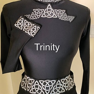 May include: Black dance costume with silver Celtic knot embroidery. The costume includes a collar, a chest piece, a wrist cuff, and a belt. The word "Trinity" is displayed in white text on the front of the costume. The costume is designed for dance.