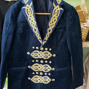 May include: A navy blue velvet blazer with gold and blue Celtic knot embroidery on the lapels and front. The jacket has a classic tailored design and is suspended from a wooden hanger.