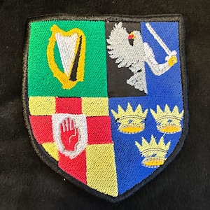 May include: A shield-shaped embroidered patch featuring four quadrants. The top left is green with a yellow harp. The top right is blue with a silver bird holding a sword. The bottom left is a red hand on a yellow and red background. The bottom right is blue with three yellow crowns.